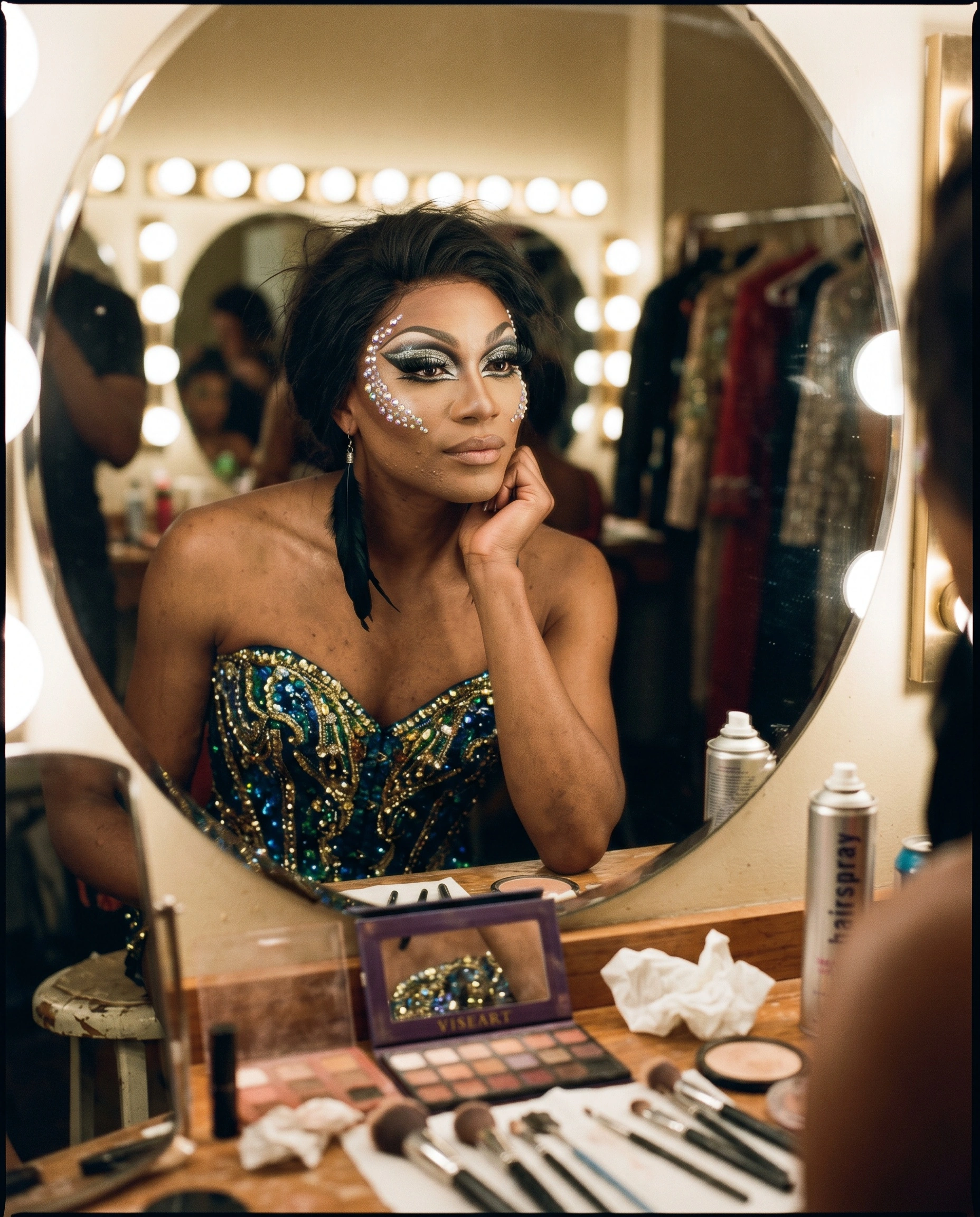 Reflected in a vanity mirror ringed with warm bulbs, a drag performer leans chin to hand in a jewel-encrusted teal-and-gold bustier, pearl-flecked eye makeup gleaming. Spent makeup brushes, hairspray cans and tissues litter the counter, a rack of sequined gowns blurs behind. Amber tungsten, candid backstage intimacy, medium-format grain.