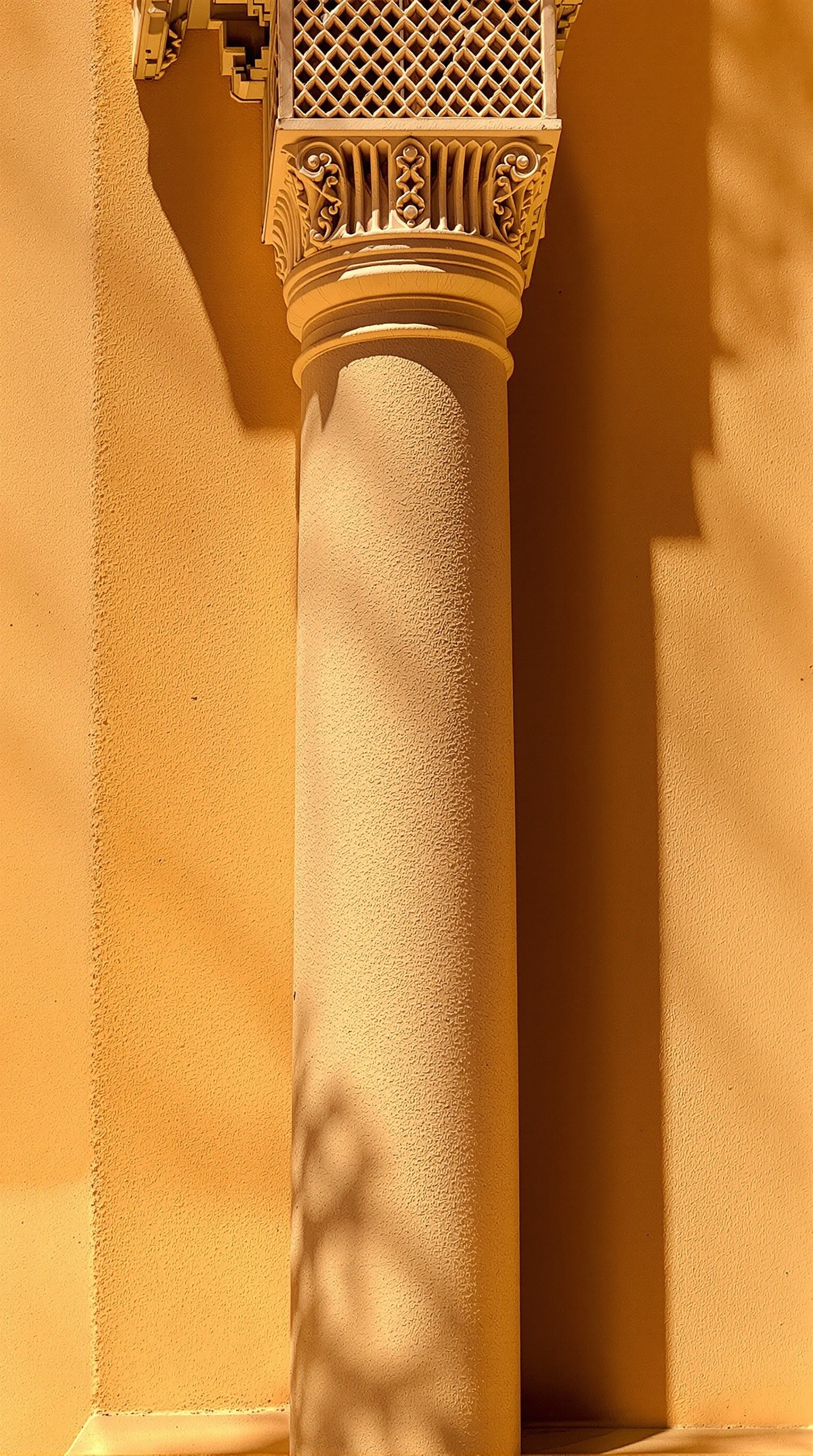 Honey-coloured plaster column rising against a sun-baked ochre wall, its filigreed capital throwing crisp geometric shadow across the surface. Hard noon light bleaches one face into pale buttercream while the other stays burnt amber. Saturated golden palette, architectural minimalism, Moroccan riad calm.