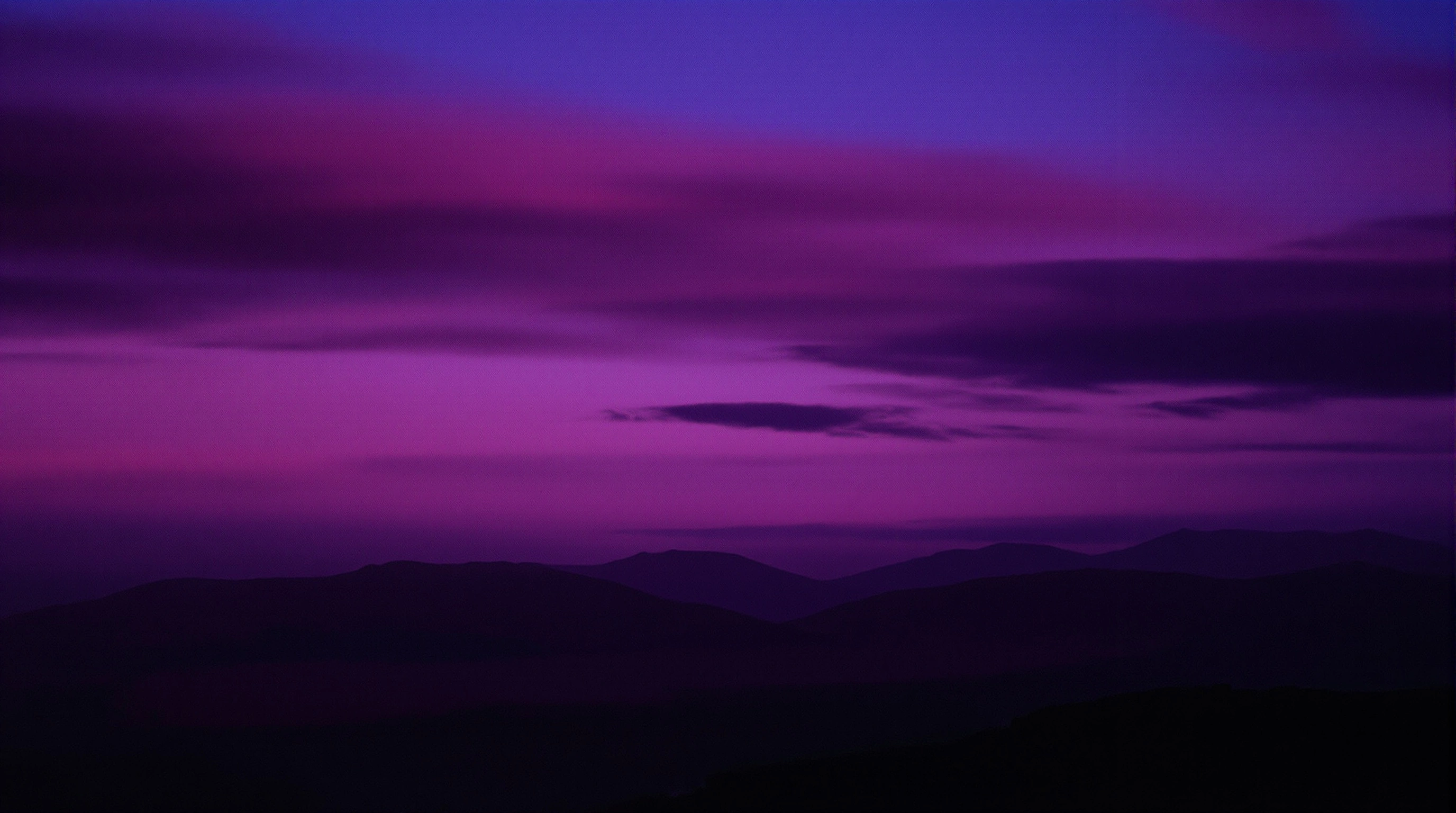 Layered bands of aubergine and magenta cloud streak a saturated post-sunset sky above the soft silhouettes of receding violet ridgelines. Faint mist clings between the hills, the foreground a near-black slope. Wide cinematic crop, painterly colour gradation, the dense velvet tonality of Velvia at twilight.