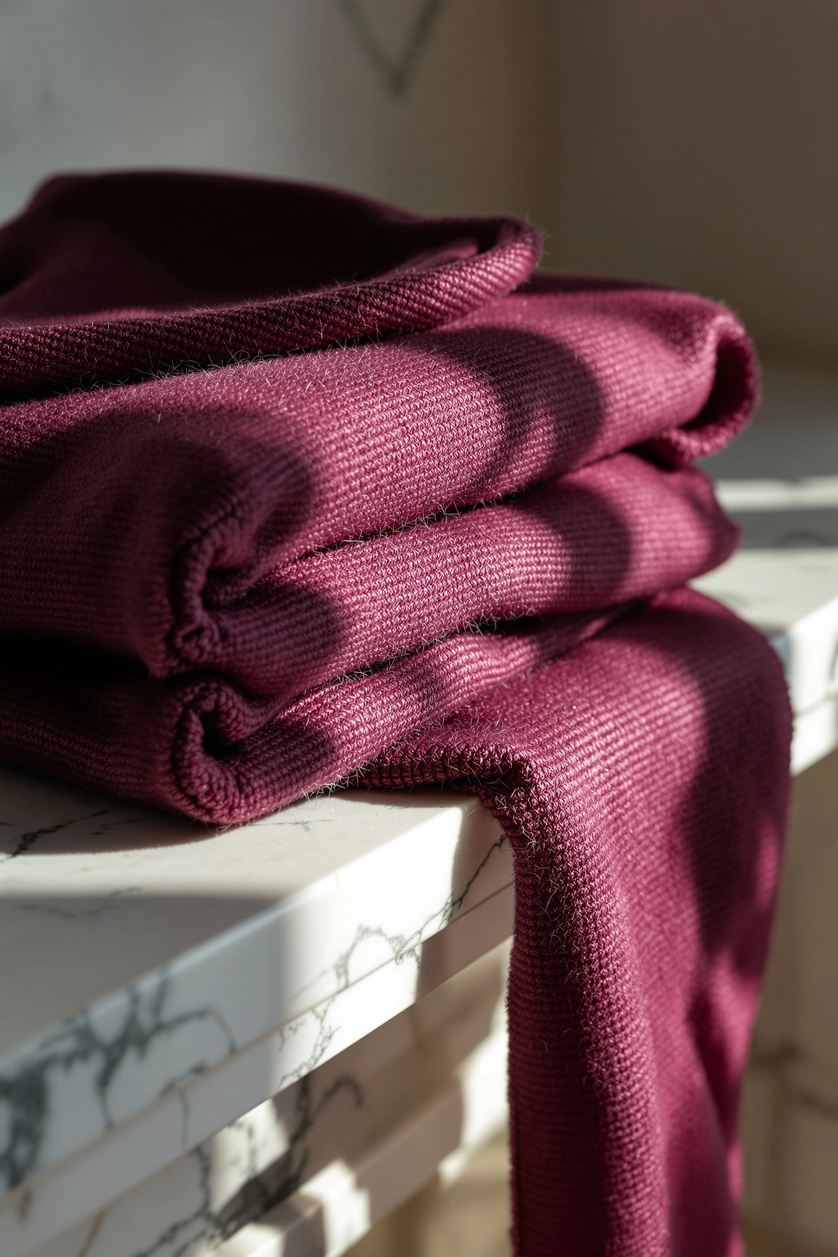 A coarse-knit wine cashmere scarf folds in thick stacked layers on the edge of a veined Calacatta marble shelf, its loose end cascading down across the cold grey stripes. Late afternoon sun rakes across the wool, igniting raised loops into magenta highlights. Tactile fashion-editorial detail, jewel and stone duet.