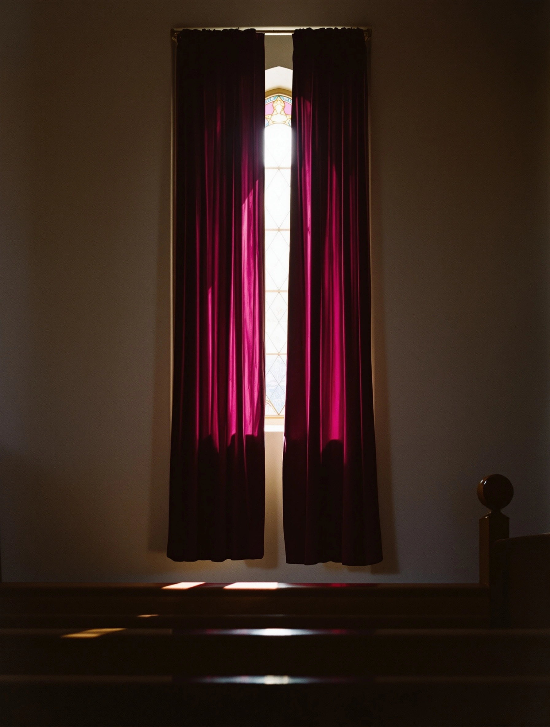 Tall magenta velvet drapes flanking a slim stained-glass window, sunlight bleeding through the gap and pooling on the chapel floor. A dark wooden pew finial sits in the foreground, taupe plaster walls absorbing the hush, reverent medium-format stillness with painterly devotional warmth.