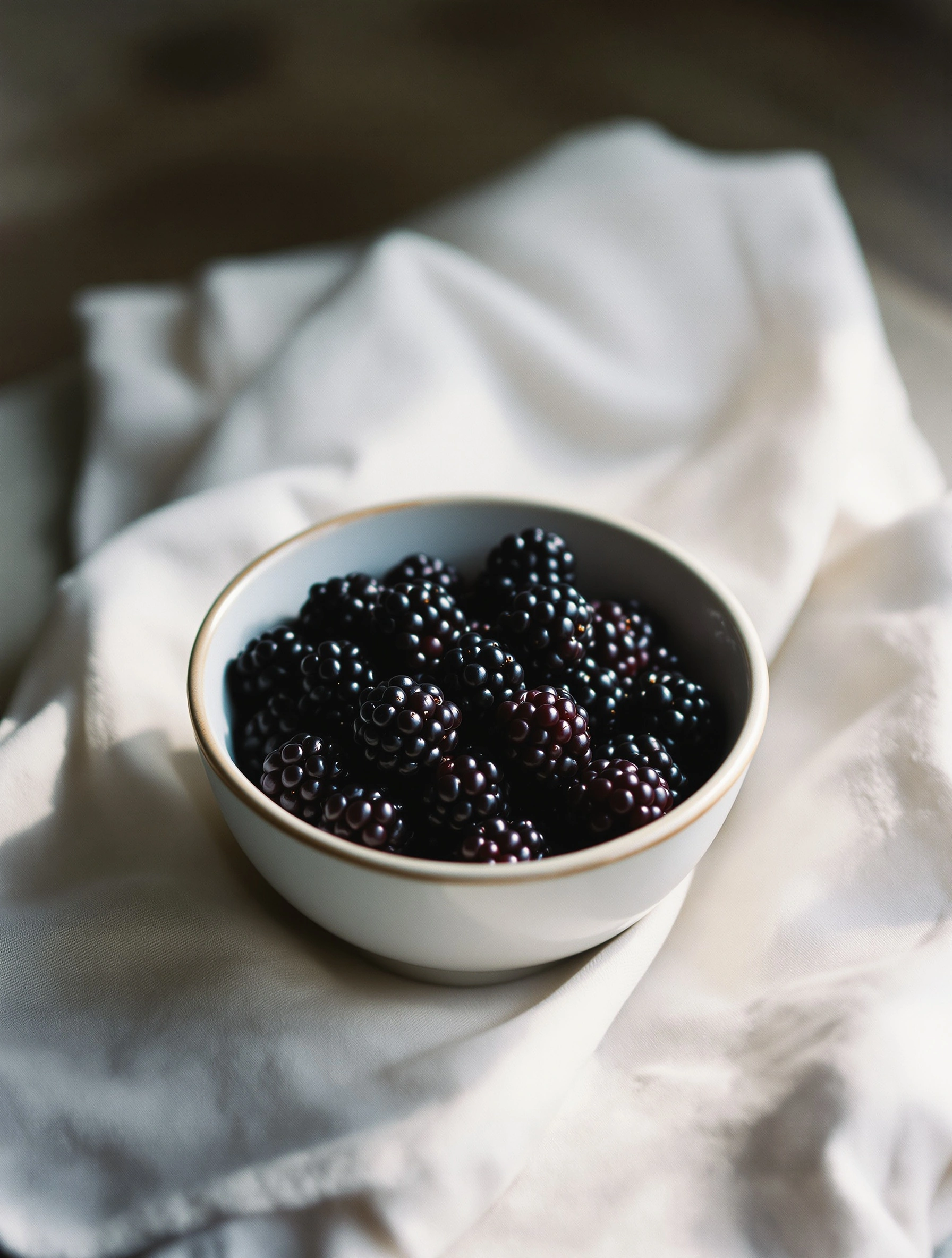 A small porcelain bowl rimmed in faint gold cradles a heap of plump blackberries on a crumpled ivory linen cloth, the dark drupelets glinting with purple-black highlights. North window light falls soft and even, the wooden surroundings sinking into espresso shadow. Slow medium-format film, painterly kitchen-table calm.