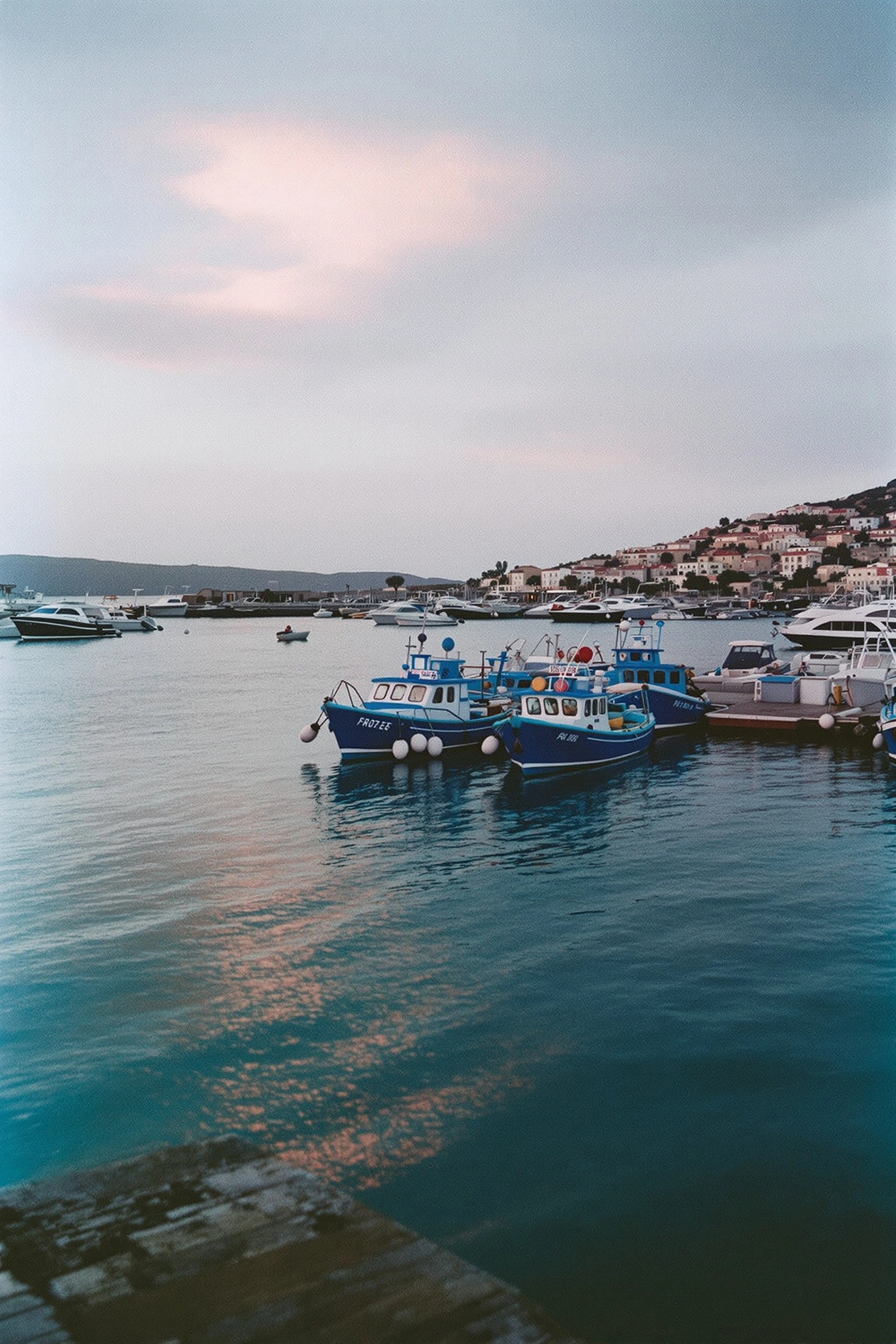 Two cobalt fishing trawlers nose against a stone quay in glassy Aegean water, white hull markings catching the last bruised pink streak of twilight. Whitewashed village lights flicker along the distant hillside while a soft mauve sky settles over still harbor reflections. 35mm film grain, Portra palette, quiet Mediterranean dusk.