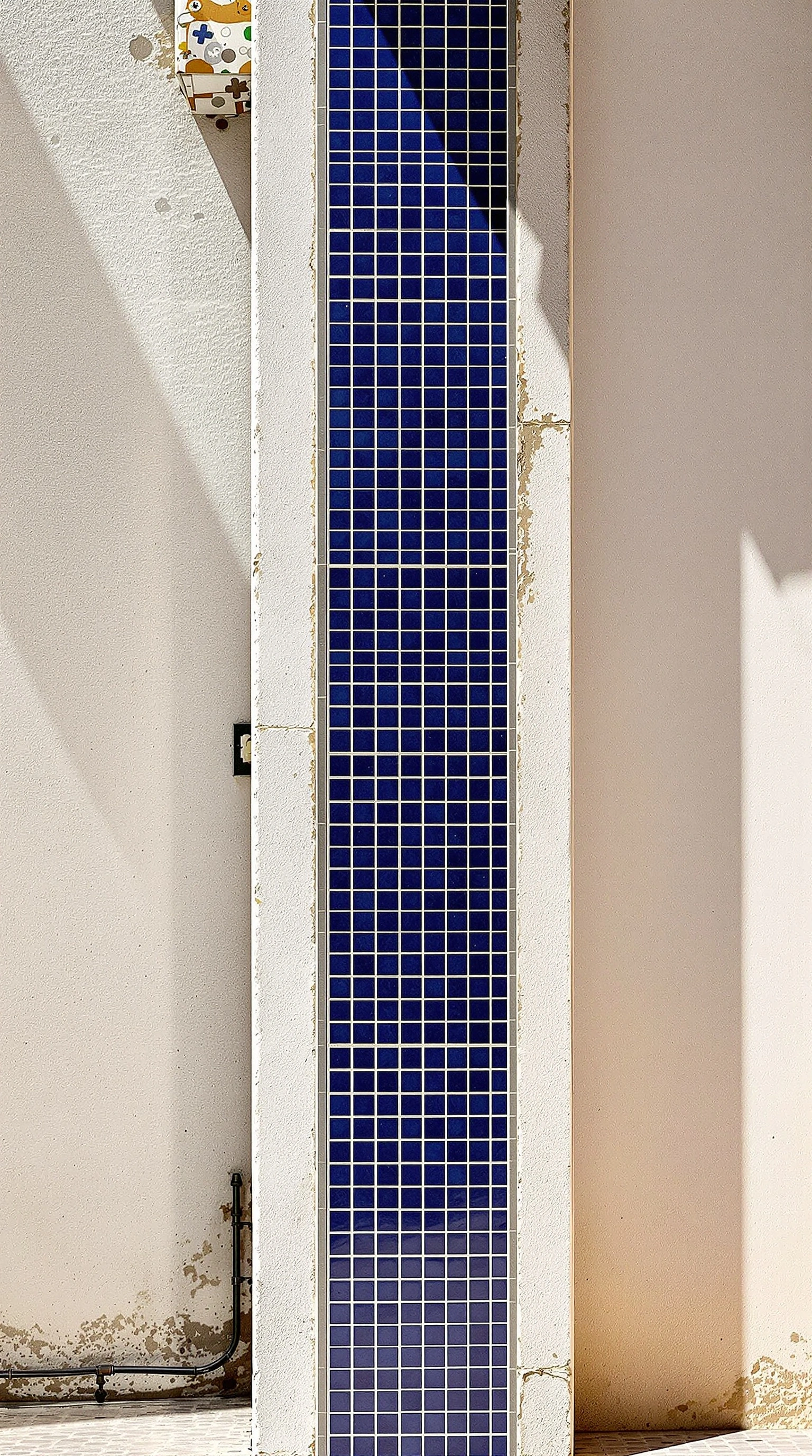 A tall slender column wrapped in cobalt mosaic tile rises through a Lisbon courtyard, flanked by chalky cream-washed stucco walls and clipped by a hard noon shadow slicing one side into deep grey. A patch of older patterned azulejos peeks from the top corner. Graphic minimal architecture, bleached light, deep blue and warm ivory color block.