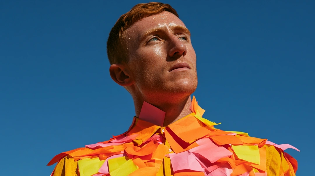 Low-angle portrait of a red-haired man gazing past the lens, neck and torso plastered in overlapping orange, yellow, and pink sticky notes flapping like scales. A cloudless cobalt sky fills the frame. Conceptual editorial sunlight, saturated complementary palette, fashion-meets-corporate satire.
