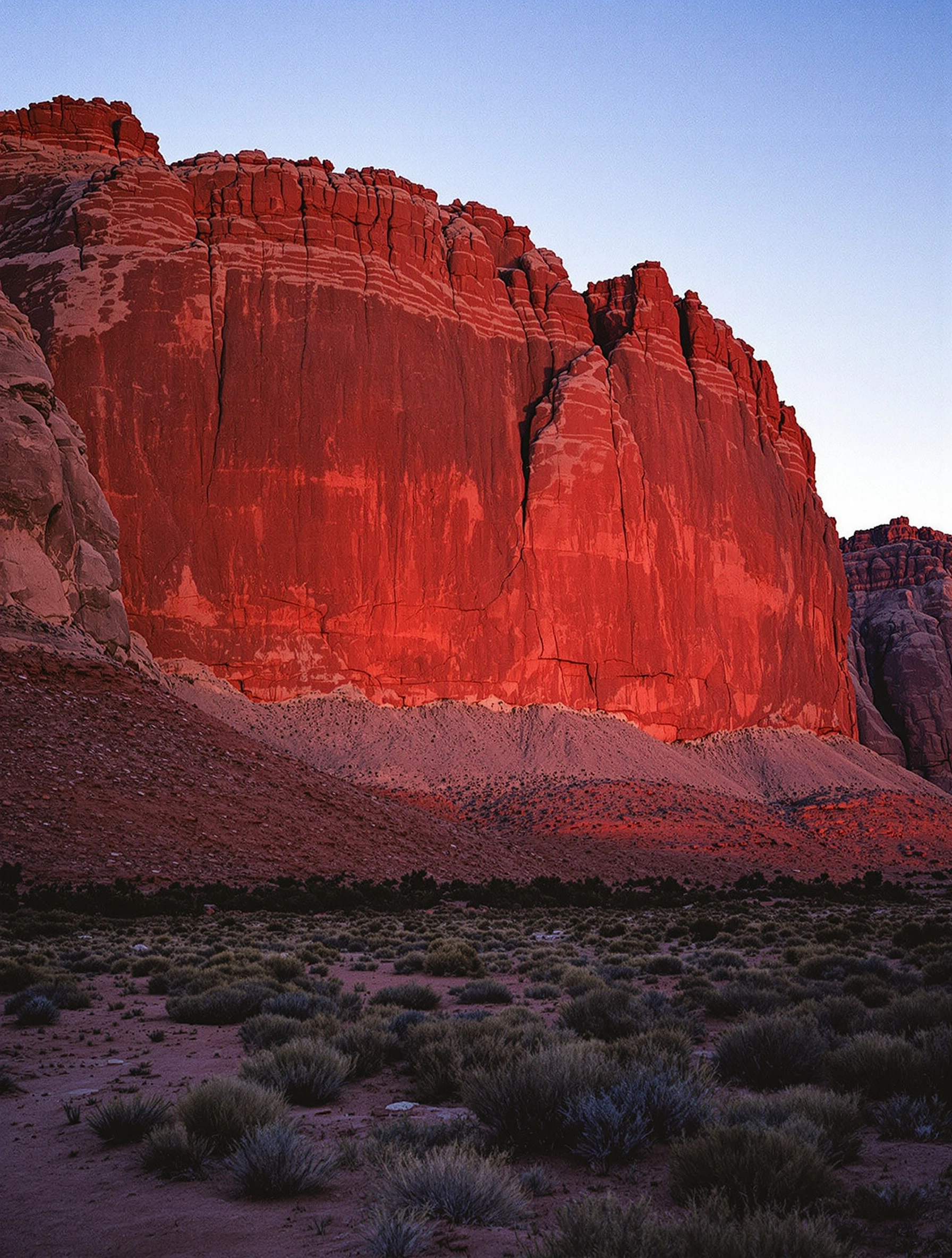 A towering sandstone canyon wall glows molten rust along its lower face while the upper buttresses fade into shaded oxblood, last sunlight catching only the western edge. Sage scrub and dusty foreground desert push depth toward the cliff, a clear cool dusk-blue sky above. Wide-angle 65mm large-format film feel, saturated sienna and turquoise complementaries.
