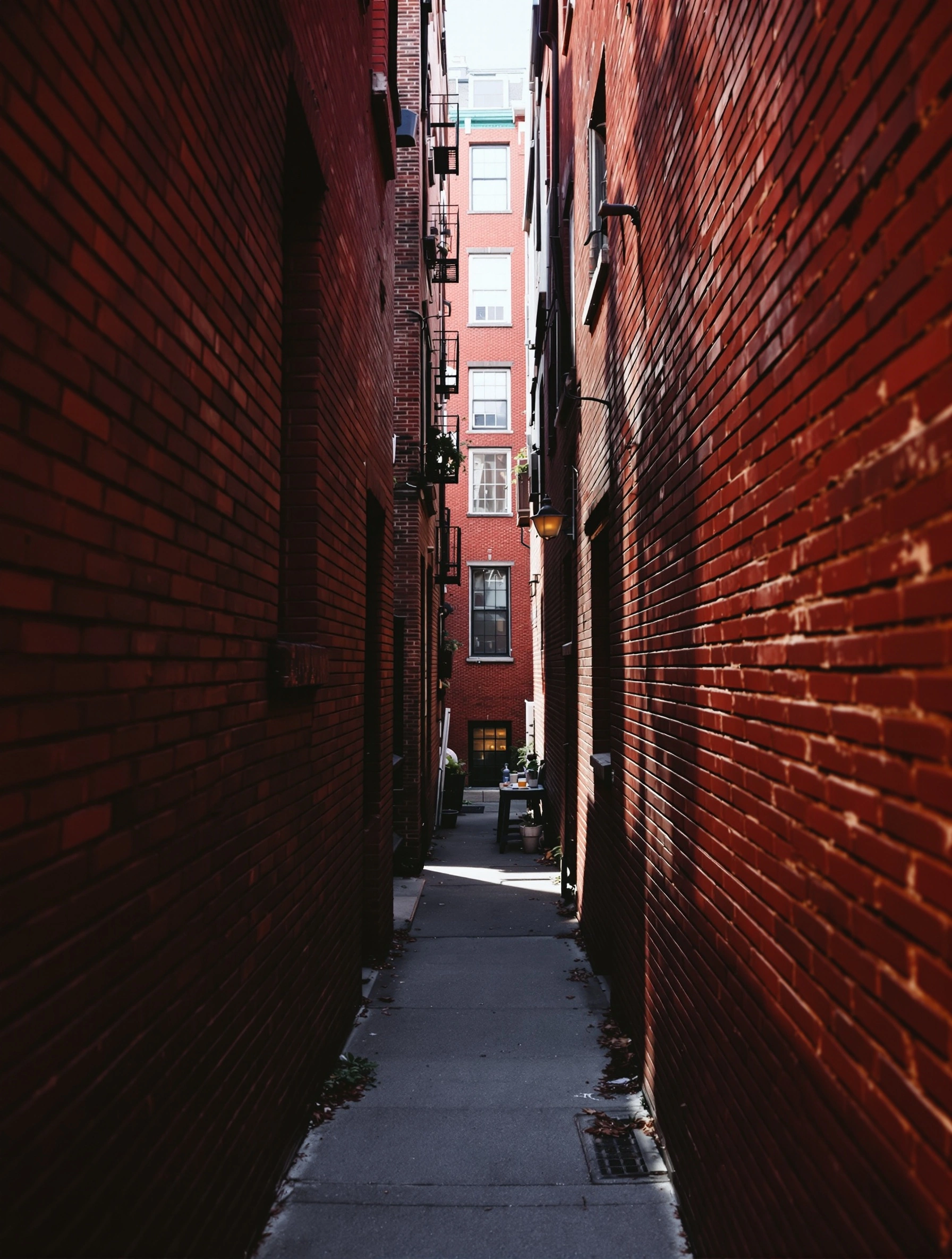 A long red-brick alley narrowing to a sunlit brownstone facade, fire escapes ladder up the right wall and a brass lantern juts from the left. Late-afternoon sun rakes one wall into hot ochre, plunging the other into deep shadow. Urban symmetry, crimson and umber palette.