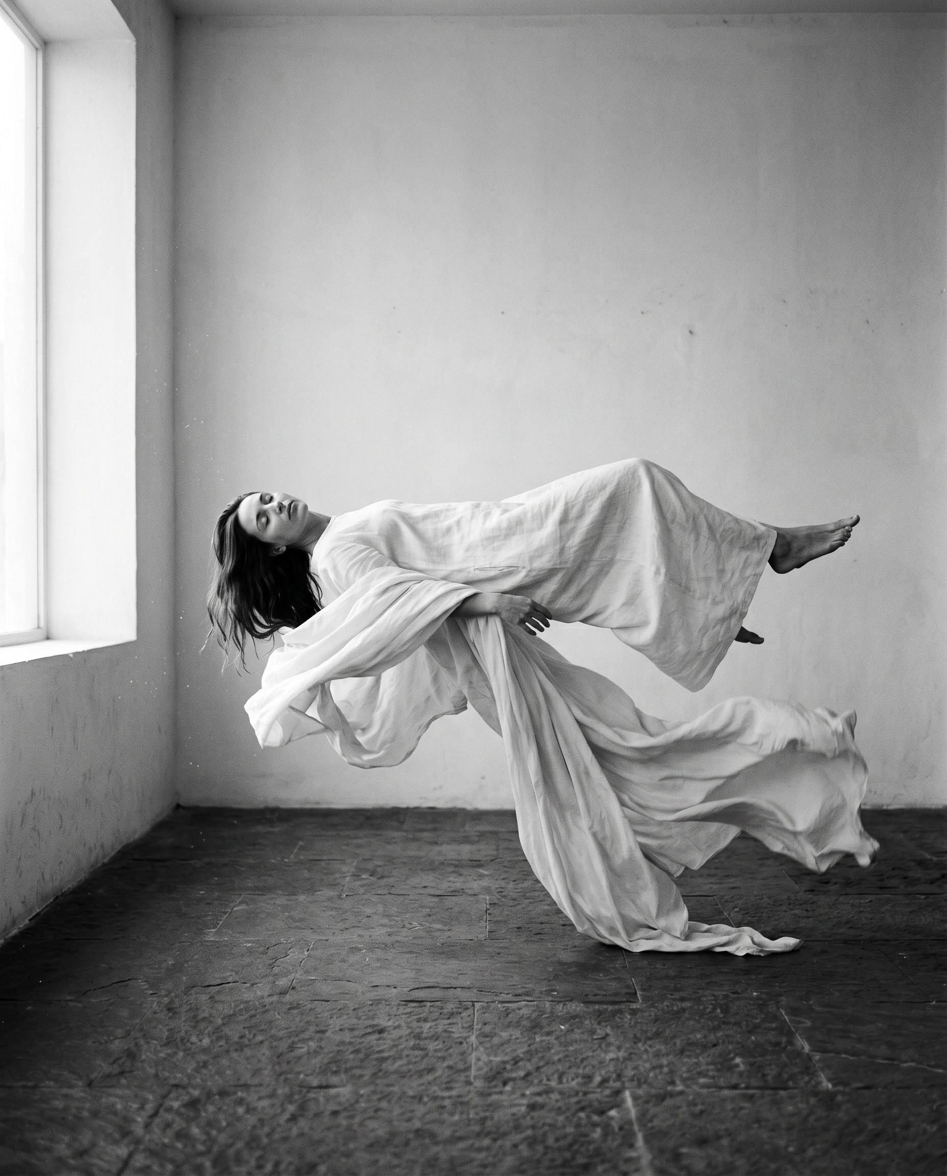 Black-and-white study of a woman in a long ivory linen gown floating horizontally above a worn stone floor, head tipped back, fabric trailing into the air behind her. A tall side window pours flat, milky daylight across a bare plaster room. Medium-format silver tones, levitation as quiet metaphor, monastic emptiness.