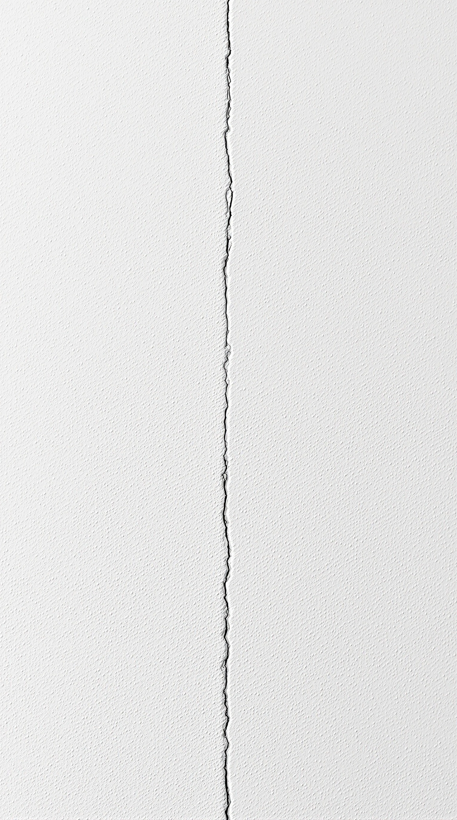 A near-empty field of textured white plaster, a single jagged hairline crack winding vertically down the center. Gentle sidelight reveals the orange-peel stipple of the surface and a barely visible vertical seam beside the fissure. Minimalist architectural abstraction, soft tonal gradient, contemplative emptiness.