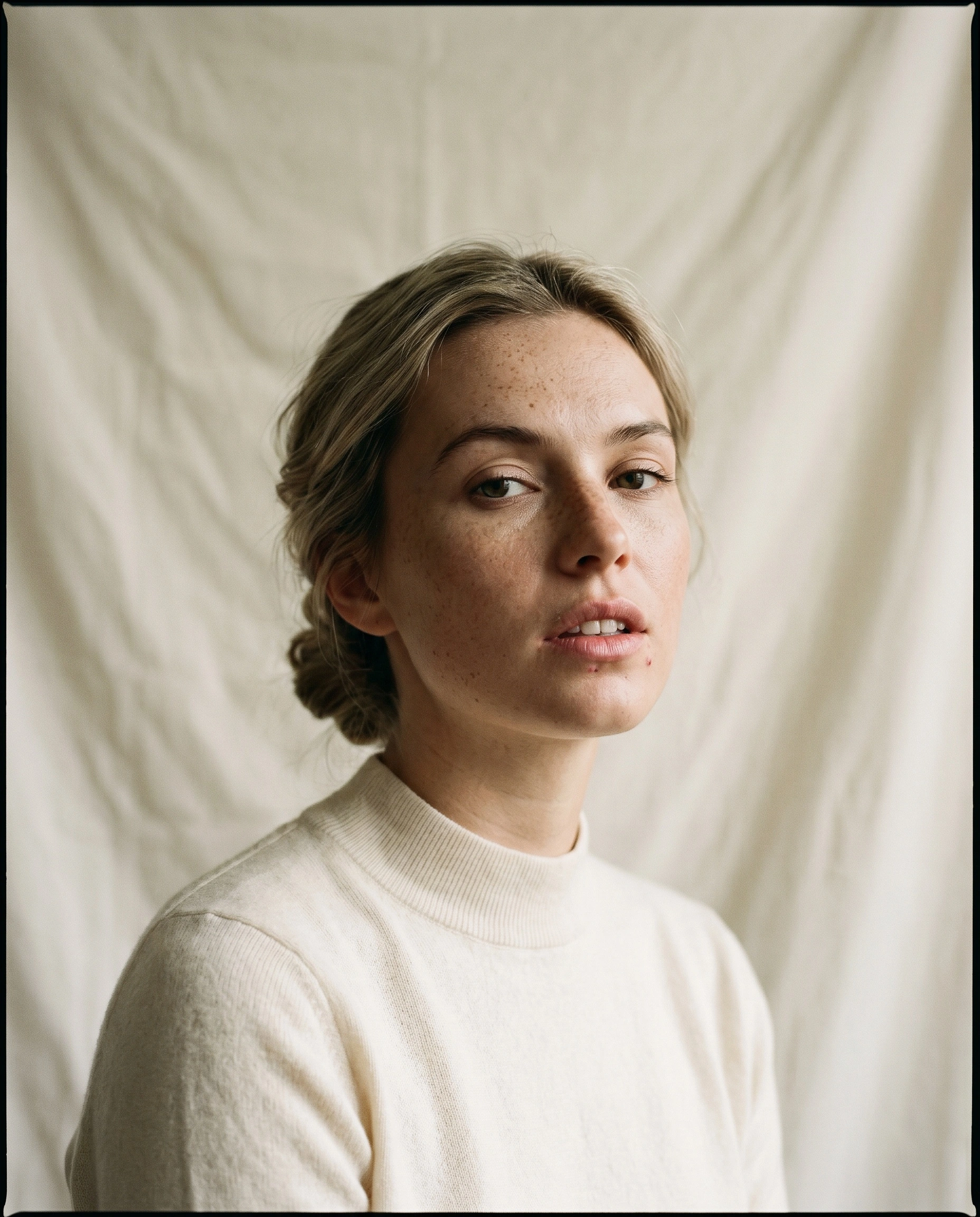 A freckled woman with ash-blonde hair pulled back in a loose braid faces the camera in a cream mock-neck sweater, lips slightly parted. She stands against a wrinkled muslin backdrop lit by diffused window light, casting gentle shadows along her jaw. Subdued ivory and skin-tone palette, medium-format portrait with the soft grain of Portra 400.