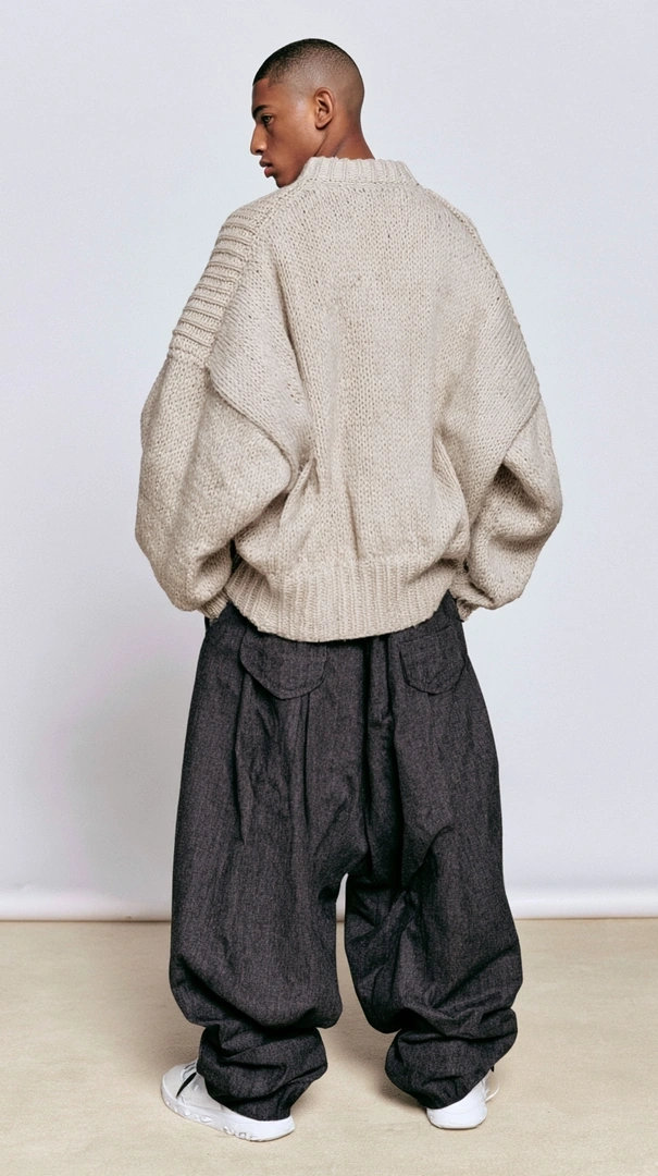 Rear-view studio shot of a model in a heavily oversized cream cable-knit sweater dropped low over slouchy charcoal pleated trousers and scuffed white sneakers. Posed flat against a seamless taupe backdrop, soft even lighting flattening every texture. Minimal fashion editorial, contemporary streetwear silhouette.