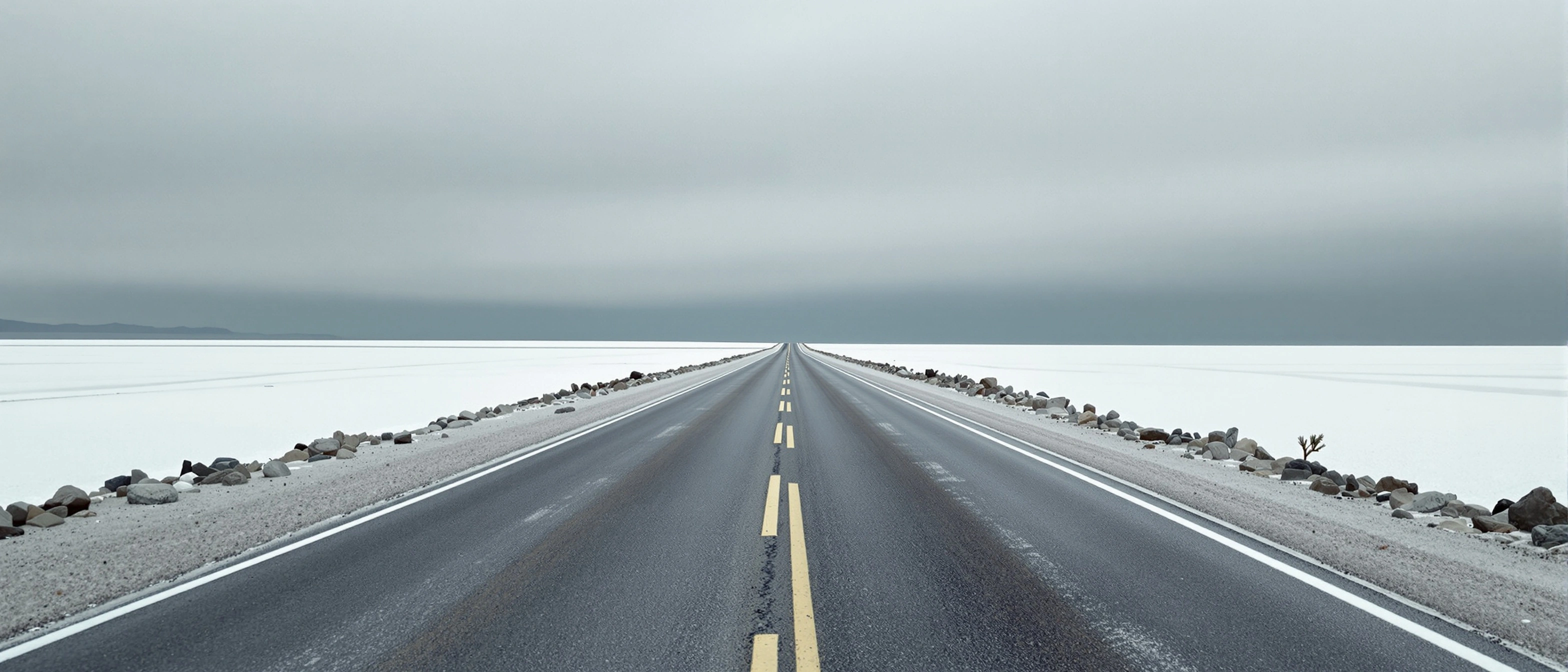 A perfectly centered black highway with worn double-yellow stripes drives to a vanishing point across pale, gravel-edged salt flats under a flat pewter sky. Faint blue hills along the left horizon, scattered roadside stones. Ultra-wide 2.66:1 frame, total stillness, the muted register of overcast medium-format colour.