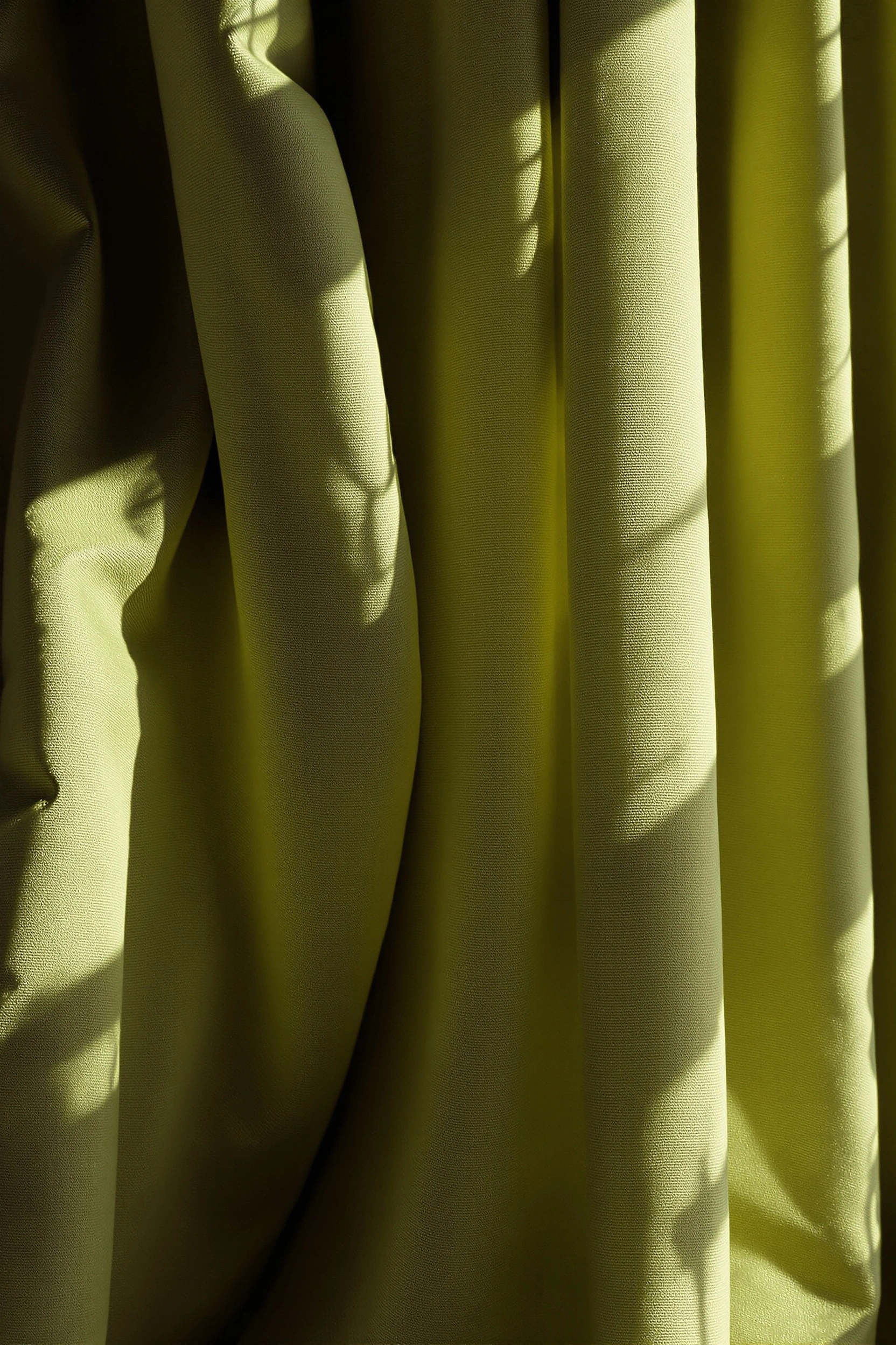 Heavy olive-sage linen drapes fall in vertical columns, raked by warm low side light that swells the pleats into soft golden ridges and deep mossy hollows. Faint window-bar patterns ghost across the weave near the top. Vertical fabric study, mid-century film palette, sculptural calm.