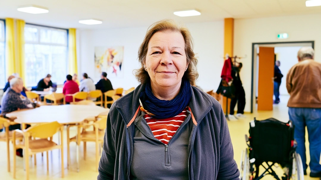 A middle-aged woman in a gray fleece zip-up and navy scarf smiles warmly toward camera inside an airy community center, yellow curtains and round tables softly defocused behind her. Bright overhead daylight, a wheelchair and seated visitors in the background. Documentary-realist tone, candid editorial warmth.
