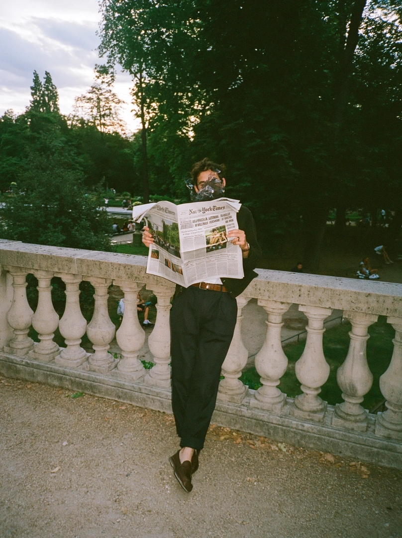 Figure in black trousers and loafers leaning against a baluster railing in a Parisian park, raising the open New York Times to obscure the face. Soft evening light filters through chestnut trees, distant strollers beyond the marble urns. 35mm film grain, romantic editorial nonchalance.