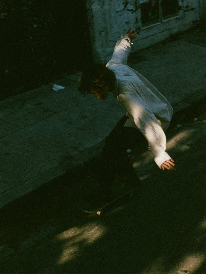 A young skater mid-kickturn on a sun-striped sidewalk, white longsleeve catching a hard slice of golden afternoon light against deep shadowed pavement. Arms flung for balance, hair in motion, ledge and weeds blurred behind. Grainy 35mm Minolta feel, high-contrast film softness, candid skate culture.