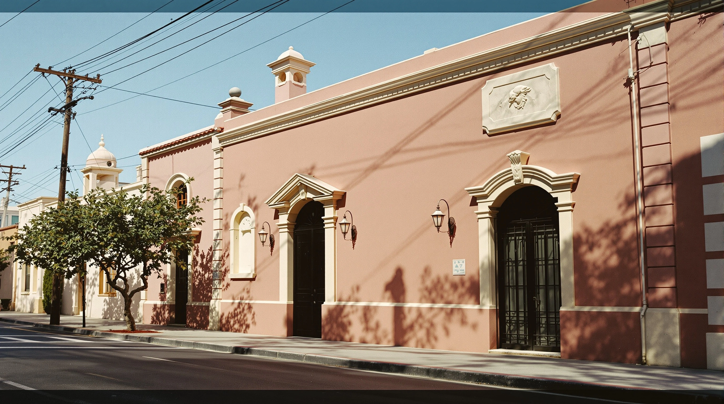 A row of dusty-rose colonial Mediterranean facades runs along an empty morning street, stucco pilasters and pedimented doorways crisply edged by low golden sun. A small citrus tree casts lacy shadows across the wall while utility wires slice a clear cobalt sky. Wide architectural frame, travel-editorial calm, lived-in pastel warmth.
