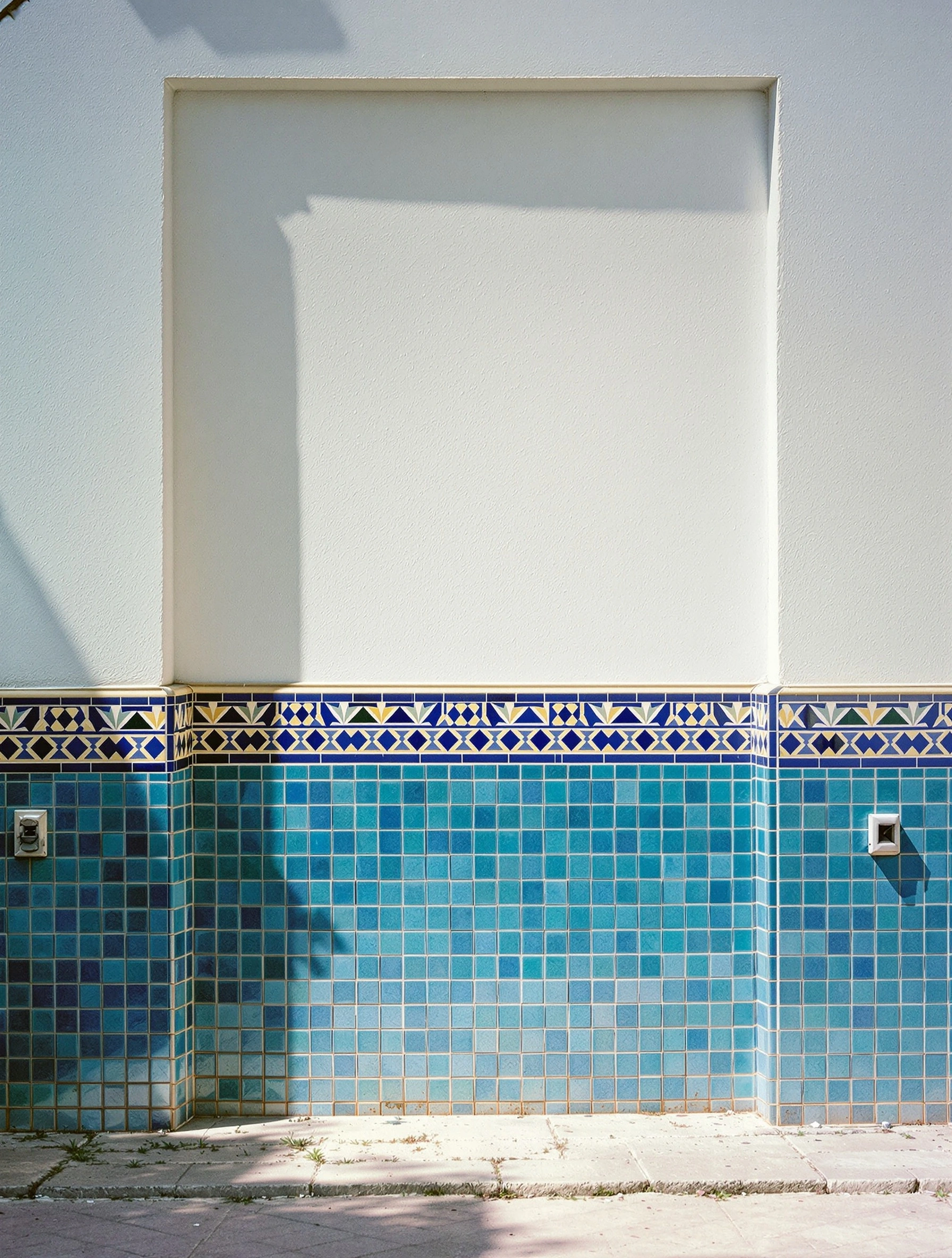 A whitewashed Moroccan exterior wall meets a dado of faded sea-glass zellige tile capped by a band of cobalt, ivory and ochre geometric border. Two small metal vents punctuate the tile field, palm shadows tracing the upper plaster. Architectural color-block frontality, hard sun, travelogue formality.