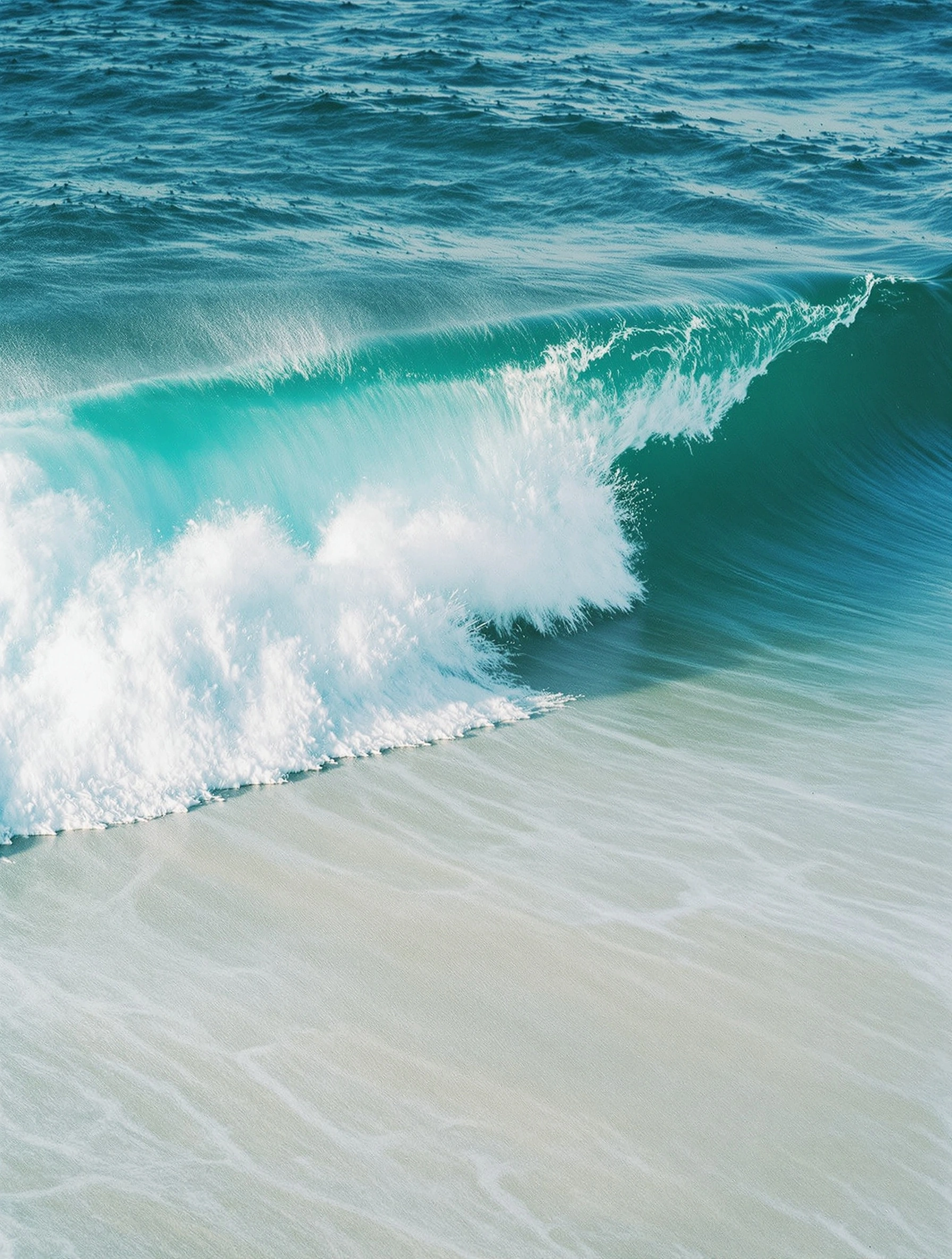 A small turquoise wave curls and detonates into white spray over a bone-pale shoreline, individual droplets pin-sharp in flight. Beyond the break, the deeper aqua sea ripples with a thousand glassy facets. Frozen ocean abstract, telephoto compression, oversaturated coastal pastels.