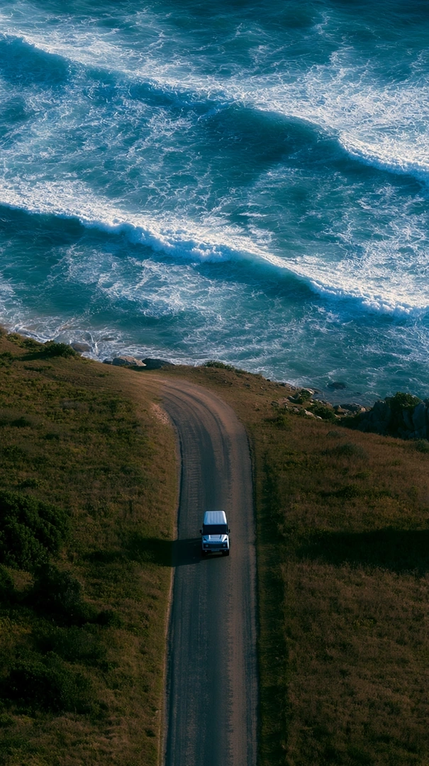 Aerial tracking view of a lone white van climbing a narrow dirt road along a green coastal headland, churning teal surf rolling onto the cliffs above. Late sunlight rakes the grass amber, vertical drone composition with road-movie isolation and sea-glass blue saturation.