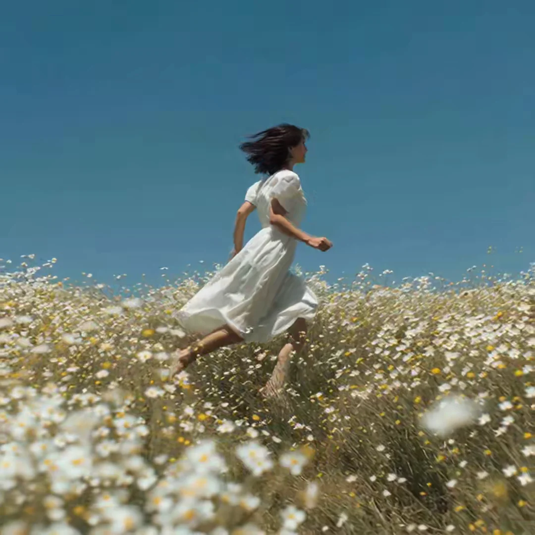 A woman in a flowing white dress leaps mid-stride through a sun-soaked meadow of daisies under a deep cobalt sky. Hair lifted in the wind, motion-blur skirt, joyful summer-film energy. Motion: she runs through the field, dress billows, daisies sway, slow-motion sun flare drifts across.