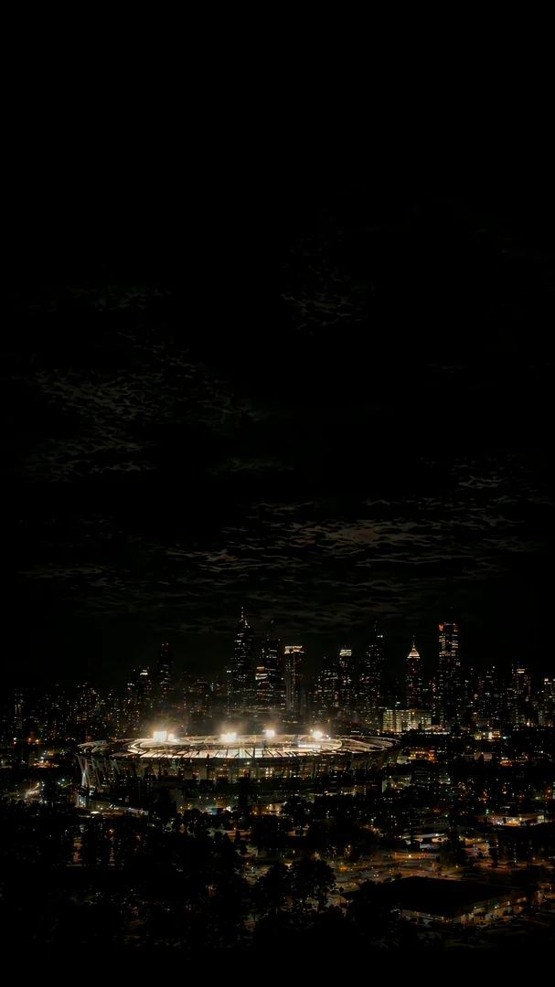 Aerial nightscape of a brightly lit stadium glowing in front of a sprawling city skyline. Cloud-shrouded black sky dominates the upper frame, warm street and stadium lights twinkling below. Wide cinematic establishing shot. Motion: slow drone push-in, stadium lights flicker, distant traffic glows, clouds drift overhead.