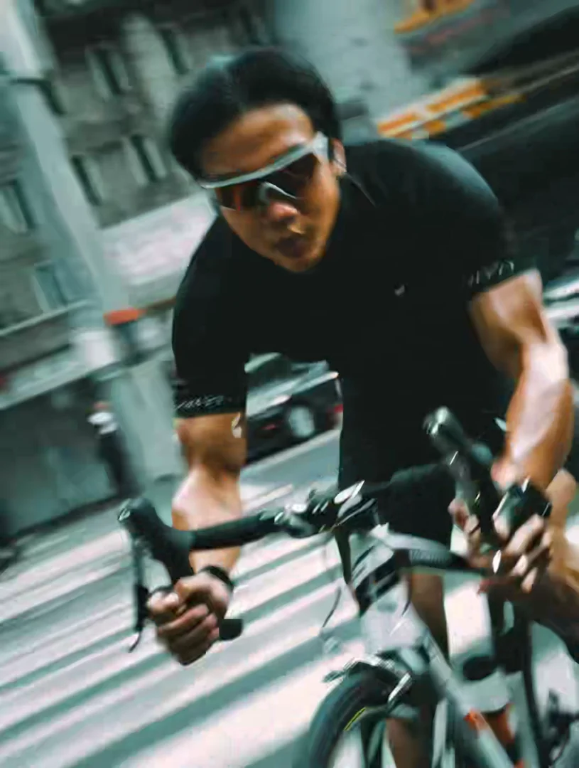 Low-angle action shot of an Asian cyclist in sport shades and a black jersey hammering down a city street, motion blur smearing surrounding buildings into pale streaks. Hand-held kinetic energy, sweat-and-sun realism. Motion: handlebars rock side-to-side, buildings blur past, breath puffs, gimbal bobs with stride.
