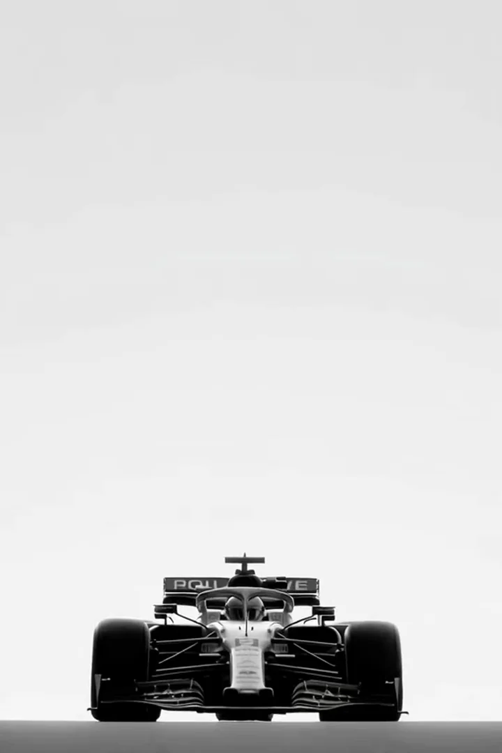 Minimalist black-and-white shot of a Formula 1 car parked head-on against a stark white void. Front wing, halo and tires razor-sharp, the rest dissolving into negative space. Editorial product framing. Motion: extremely slow push-in, faint heat shimmer, wheels rotate by a hair.