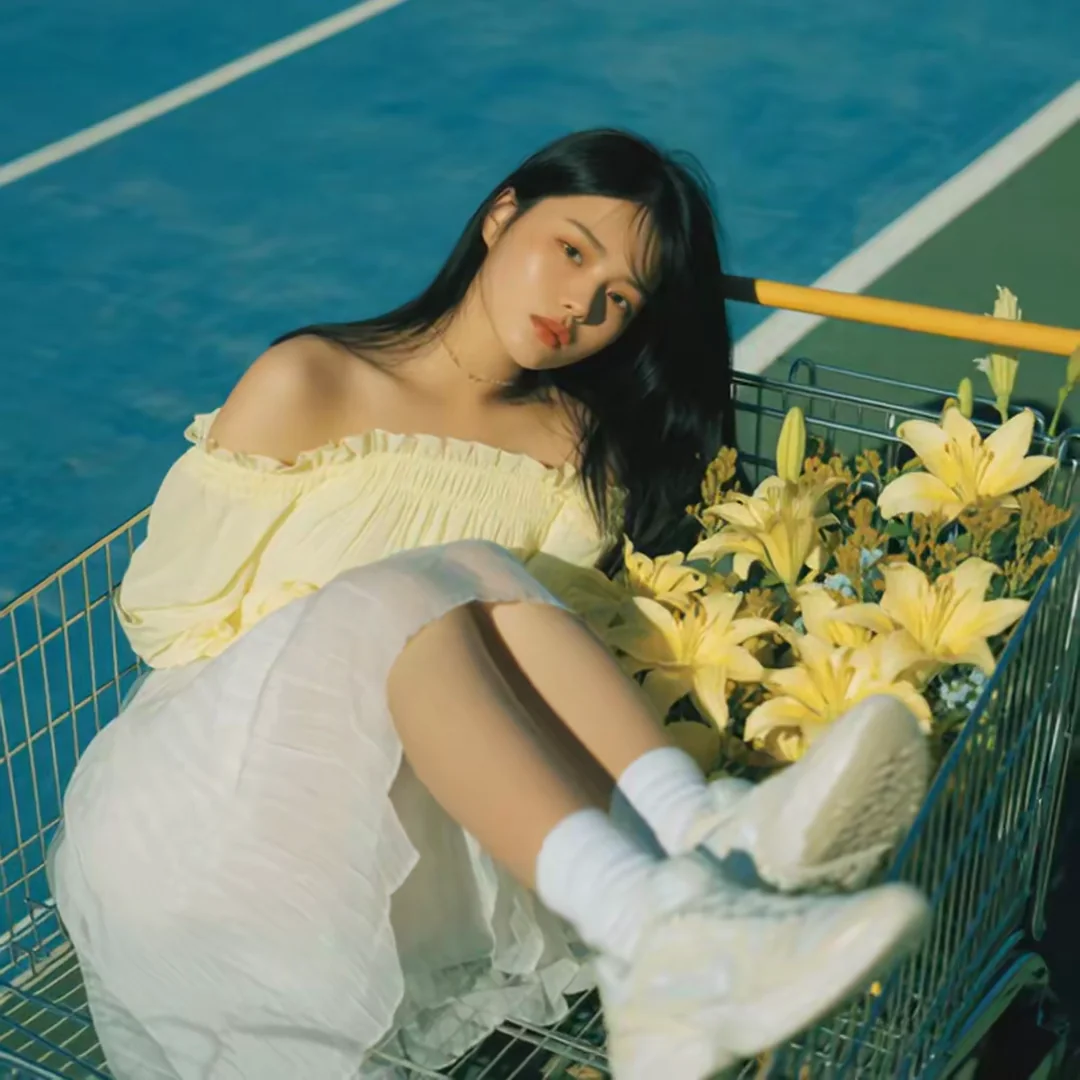 Overhead shot of a young Asian woman lounging inside a wire shopping cart on a blue running track, white dress and socks, yellow lilies piled beside her legs. Soft afternoon sun, dreamy editorial youth shoot. Motion: slow top-down drift, cart sways faintly, hair shifts, lilies tremble in the breeze.