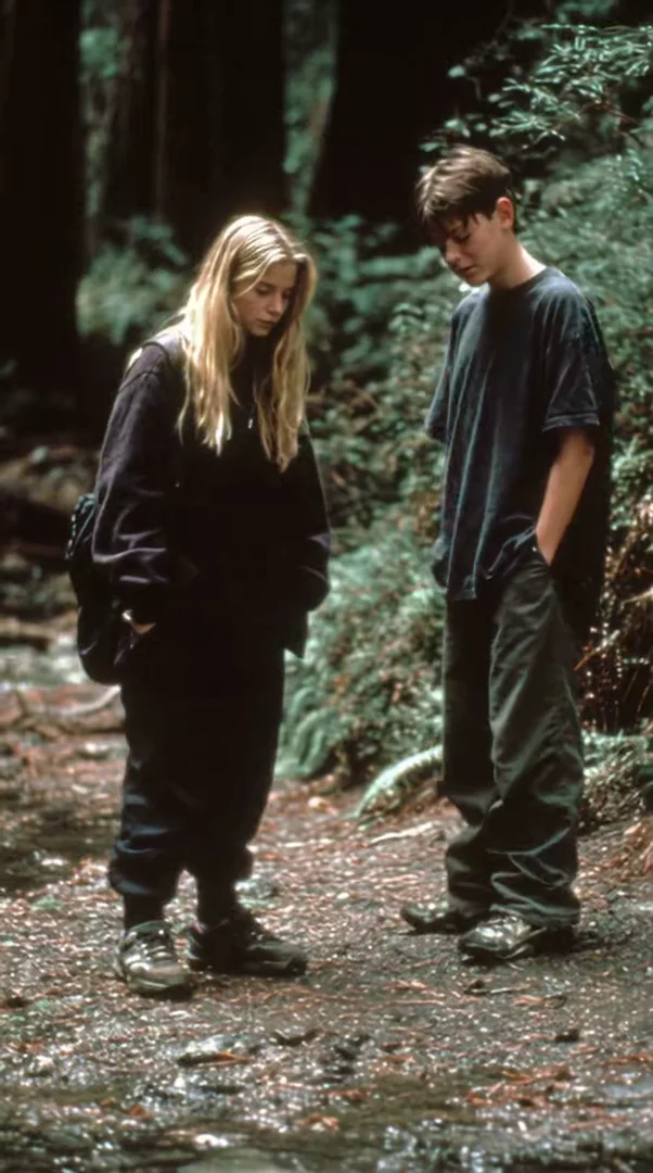 Two 90s teenagers in oversized sweaters stand on a damp redwood forest path, heads bowed in quiet conversation. Faded color-print grain, soft greens, indie coming-of-age film still. Motion: subtle handheld drift, hair shifts, fingers fidget, faint breathing.