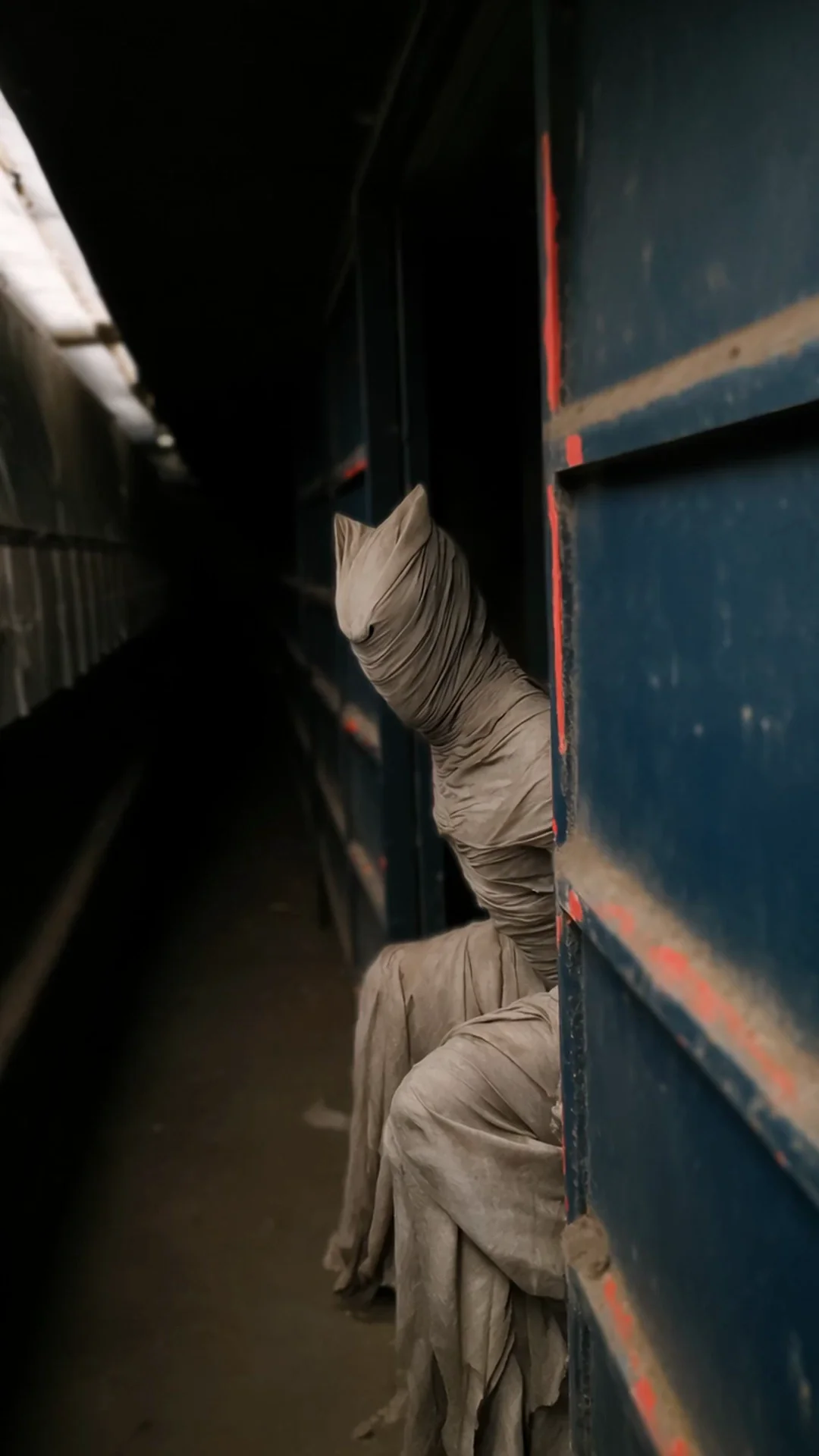 An unsettling figure entirely swathed in dusty grey bandages, head tilted in a feline silhouette, peeks from behind a rusted blue train car at a derelict platform. Hand-held analog grain, eerie cinematic dread. Motion: head tilts slowly, bandages flutter in the breeze, distant rattle, lights flicker.