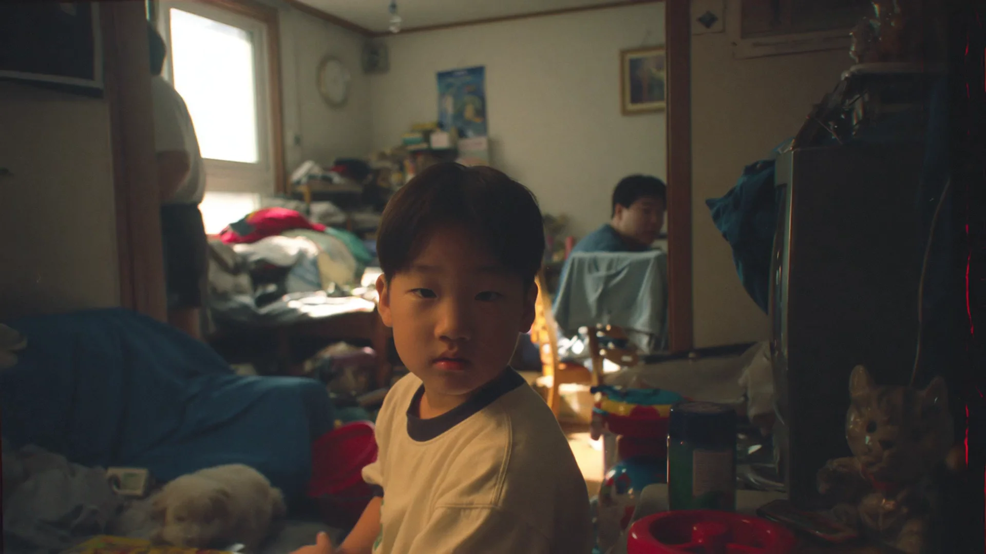 A young Korean boy in a white tee glances over his shoulder inside a cluttered family home, soft window light bathing his face. A blurred parent moves behind him. Grainy film-still warmth, intimate slice-of-life. Motion: slow push-in, boy turns his head, parent shifts in background, daylight pulses faintly.
